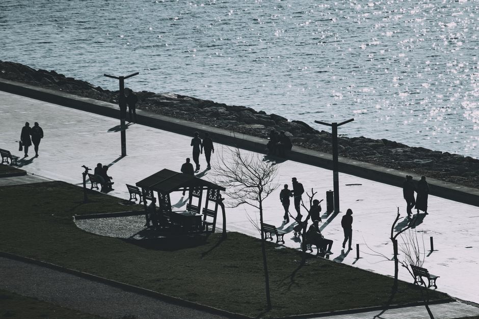Sunlit coastal promenade with silhouettes of people walking and benches along the waterfront.